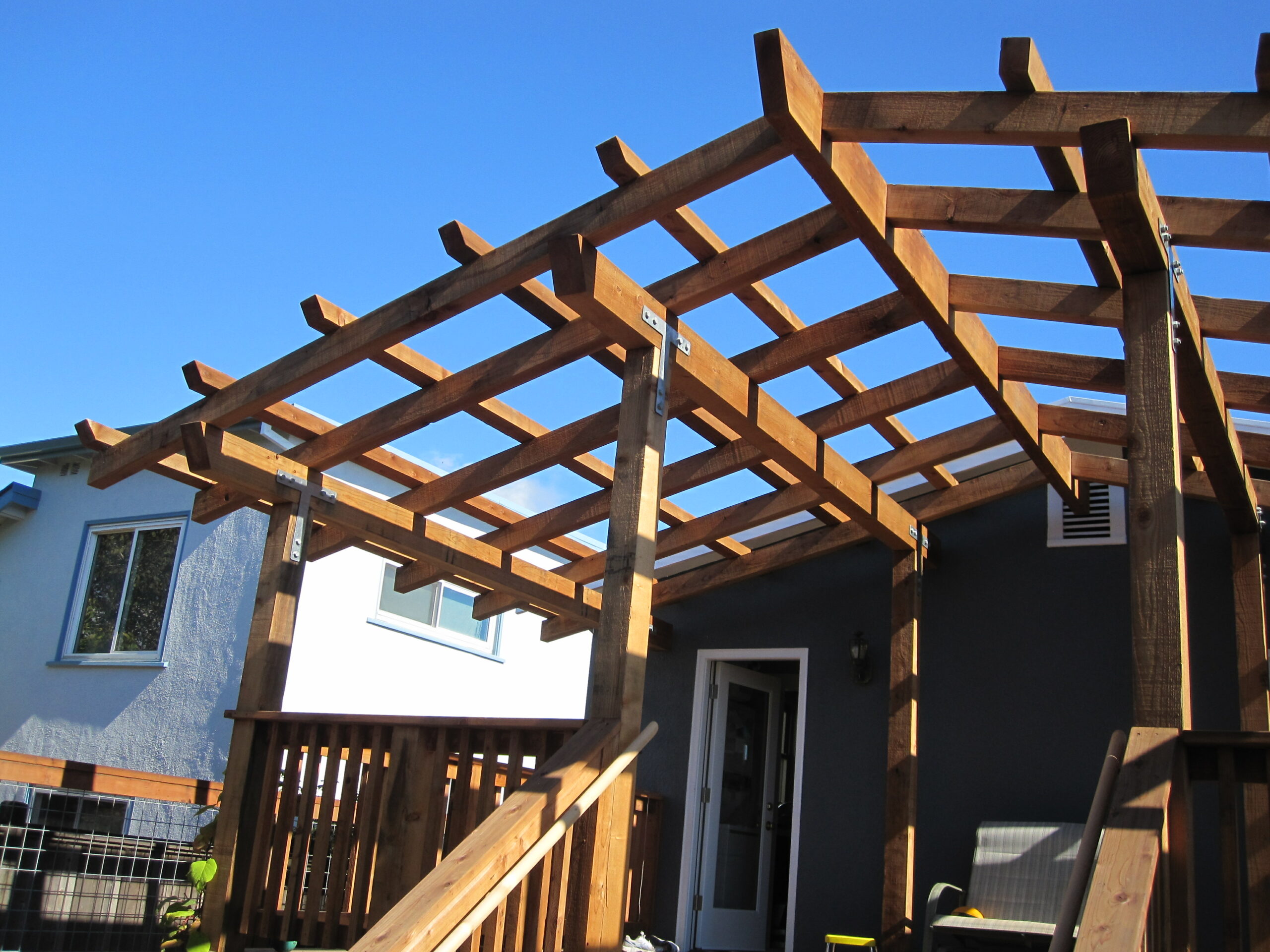 A pergola frame is structurally anchored to the rear of a house, providing coverage for the deck and staircase below.