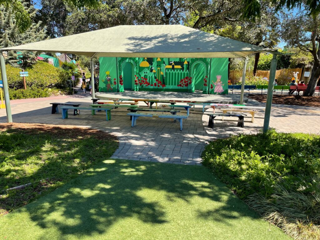 Outdoor area with built-in shade structure, adjacent open stage, mature trees, and established winding hardscape pathways.