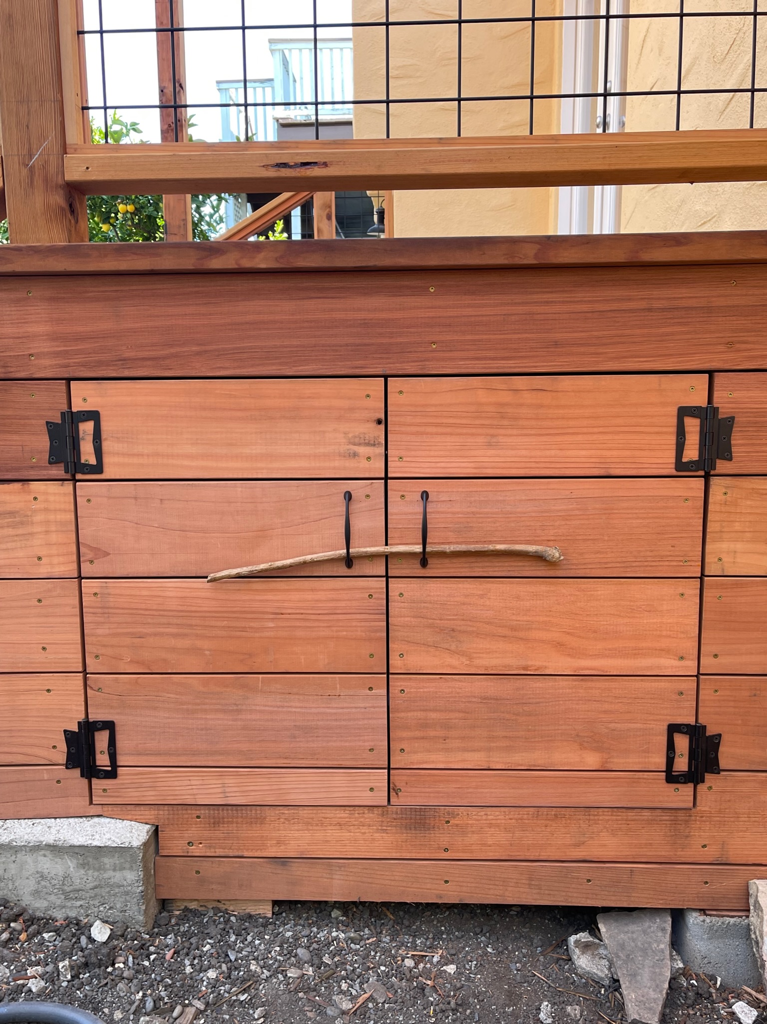 Solid wood gate with sturdy metal hardware and natural branch handle, situated on durable gravel and concrete for robust entryway use.