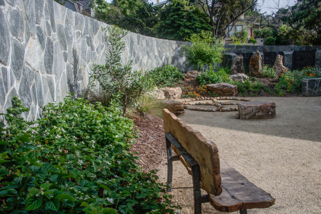 Landscaped garden with a stone fire pit, durable stone wall boundary, and established mature plantings at Northbrae Community Church.