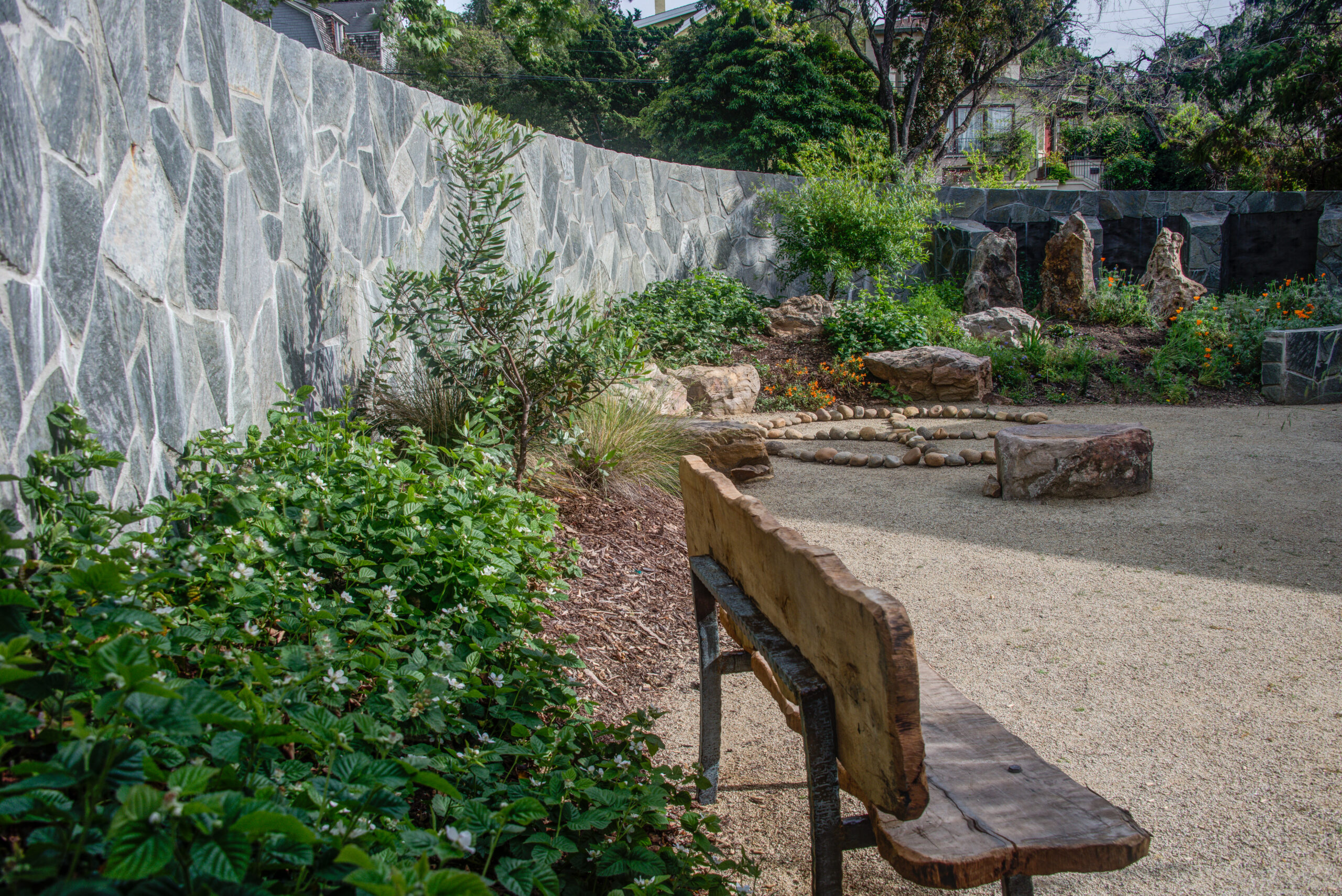 Landscaped garden with a stone fire pit, durable stone wall boundary, and established mature plantings at Northbrae Community Church.