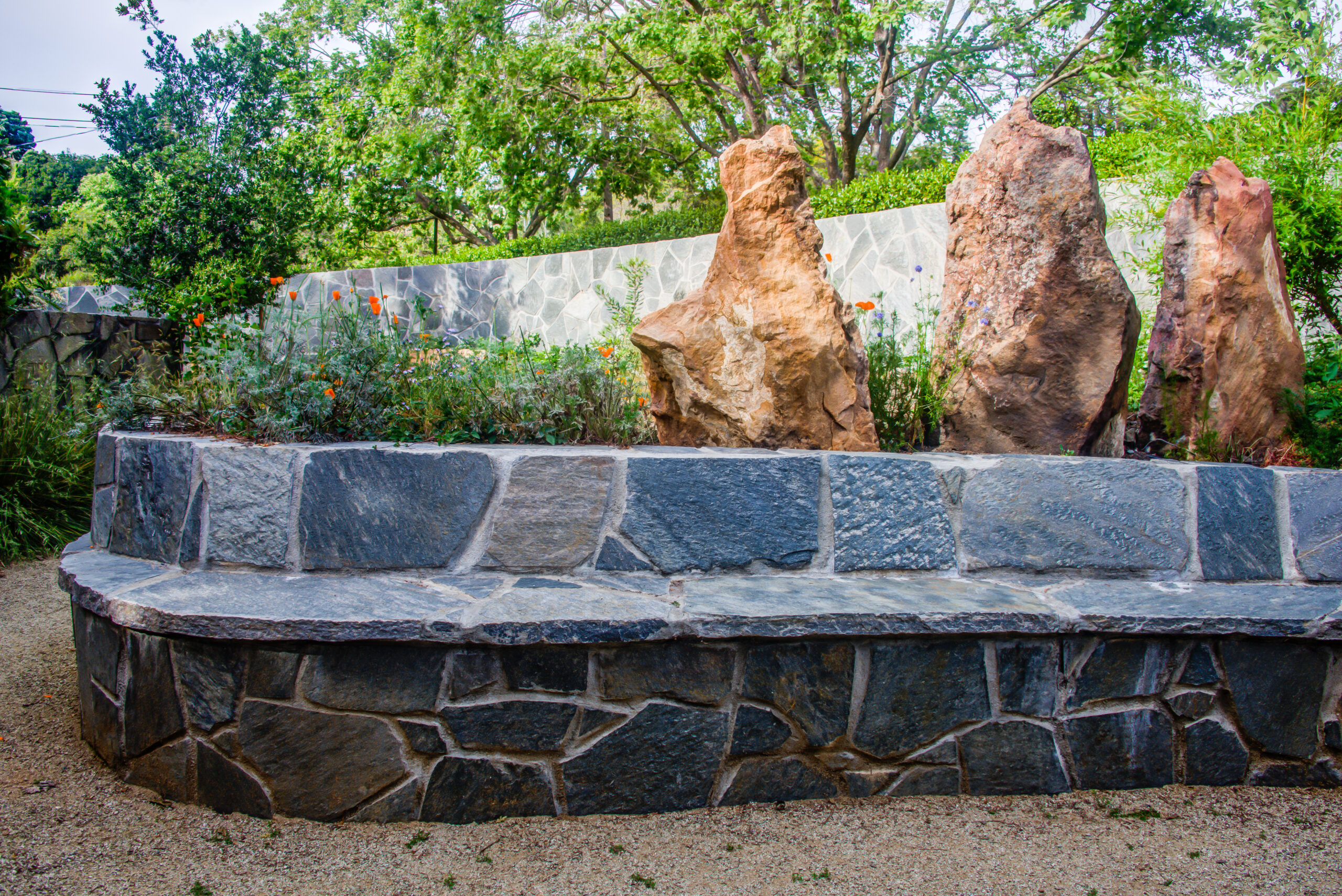 Stone bench installed before large standing rocks, with mature landscaping and existing stone wall providing privacy and structure.