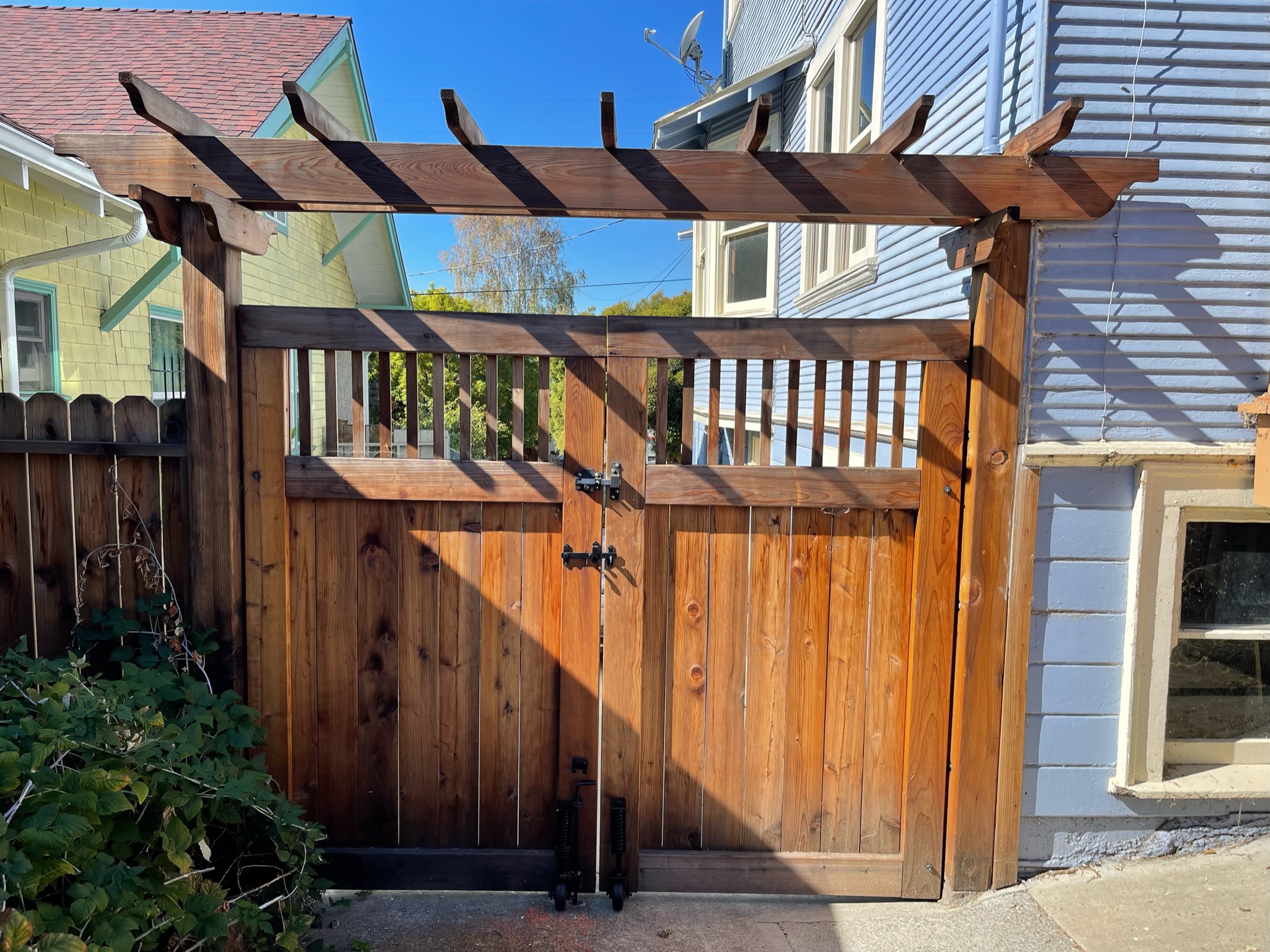 Sturdy wooden gate with integrated pergola top and durable metal hardware, positioned between two Oakland homes for privacy and security.