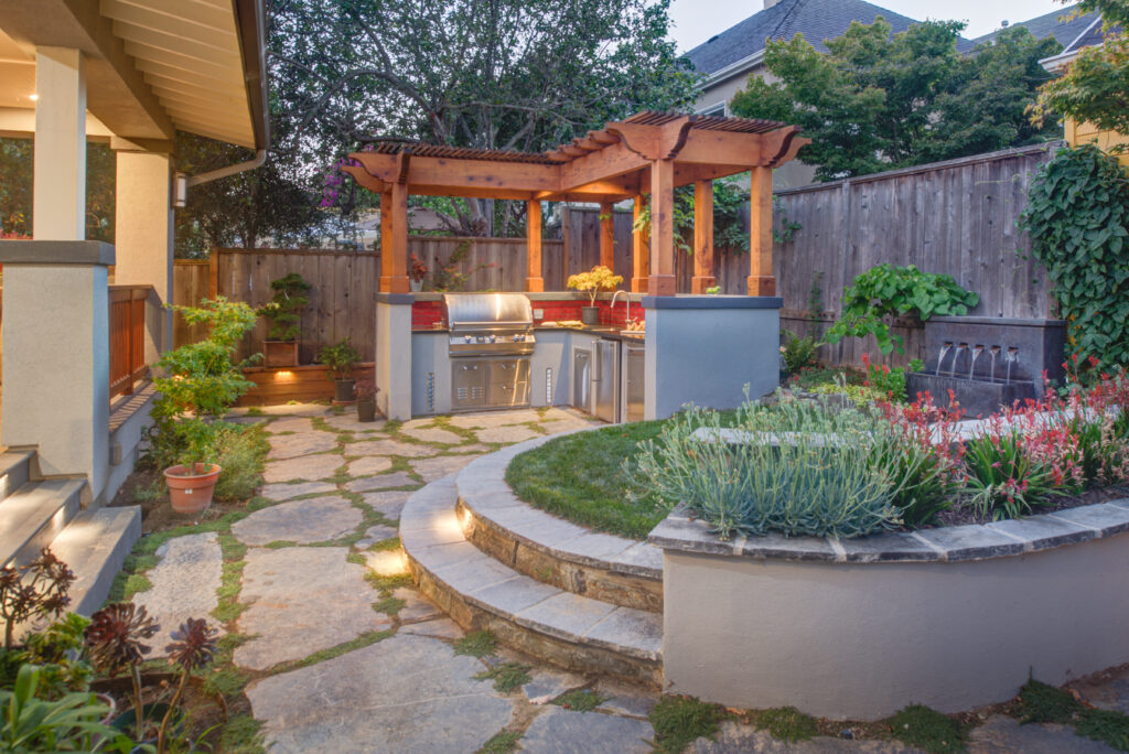 Outdoor Kitchen in Oakland with installed grill, pergola, stone patio, landscaped yard, and steps to usable fenced outdoor space.
