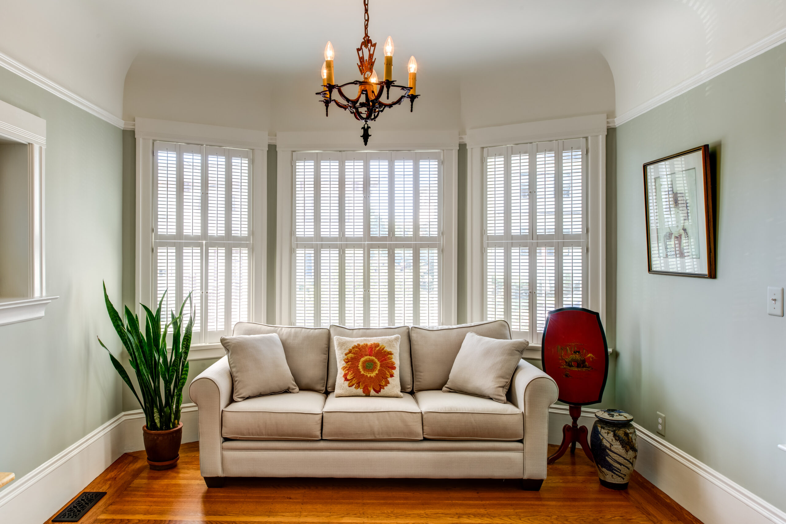 Bay windows with blinds bring natural light to a living room featuring hardwood floors and a chandelier, ideal for redevelopment.