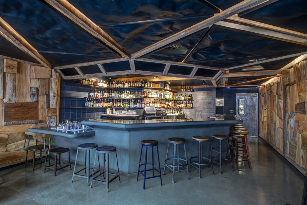 Modern bar and restaurant with open layout, exposed structural elements, concrete surfaces, and built-in shelving for storage.