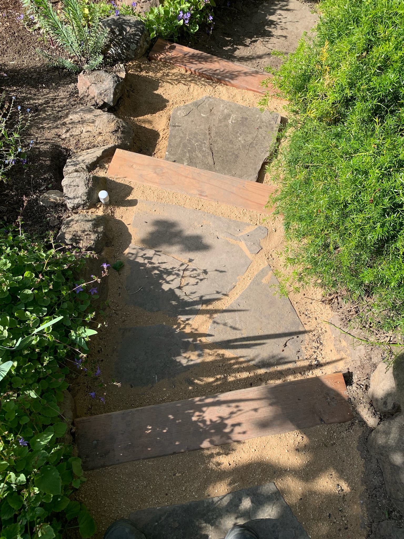 A durable stone and wood garden path with sand joints winds through backyard landscaping with mature plantings, shaded areas.