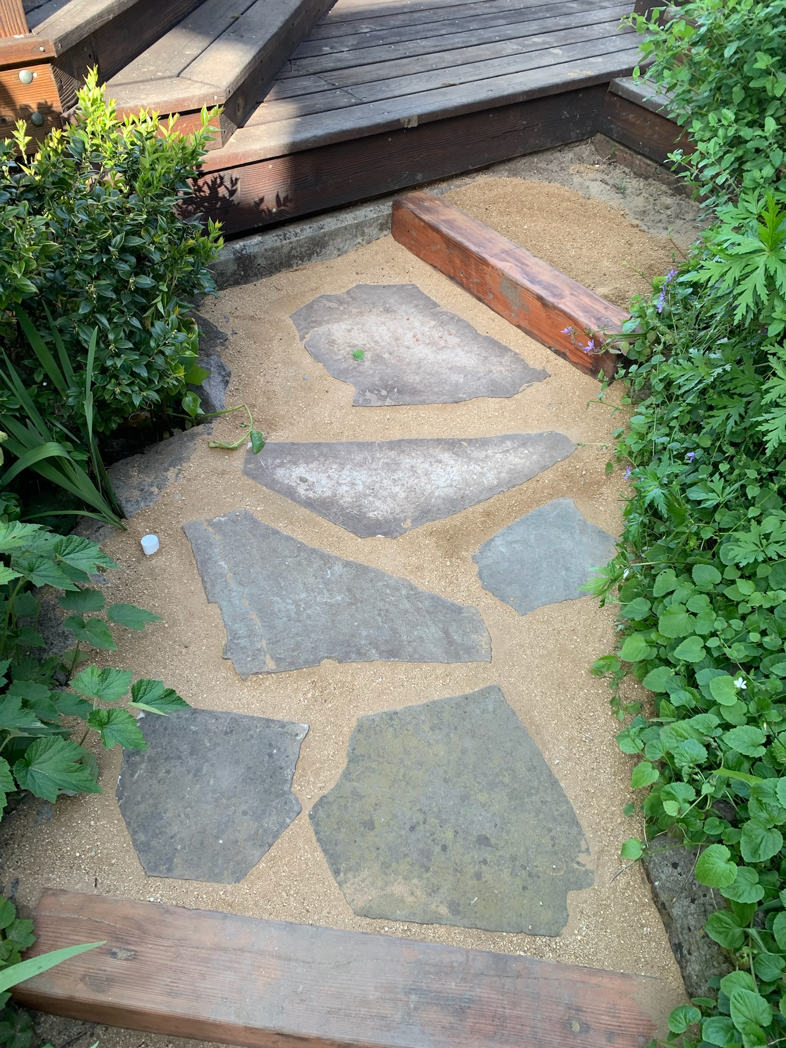 A stone pathway of large flat rocks bordered by wood beams leads to a wooden deck, showcasing durable landscape features and clear flow.