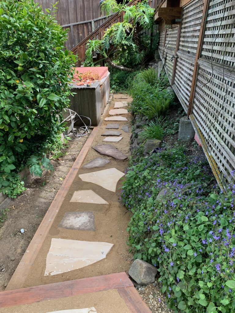 A narrow backyard garden path of irregular stone slabs runs by a wooden fence, offering potential for landscape redesign in Richmond.