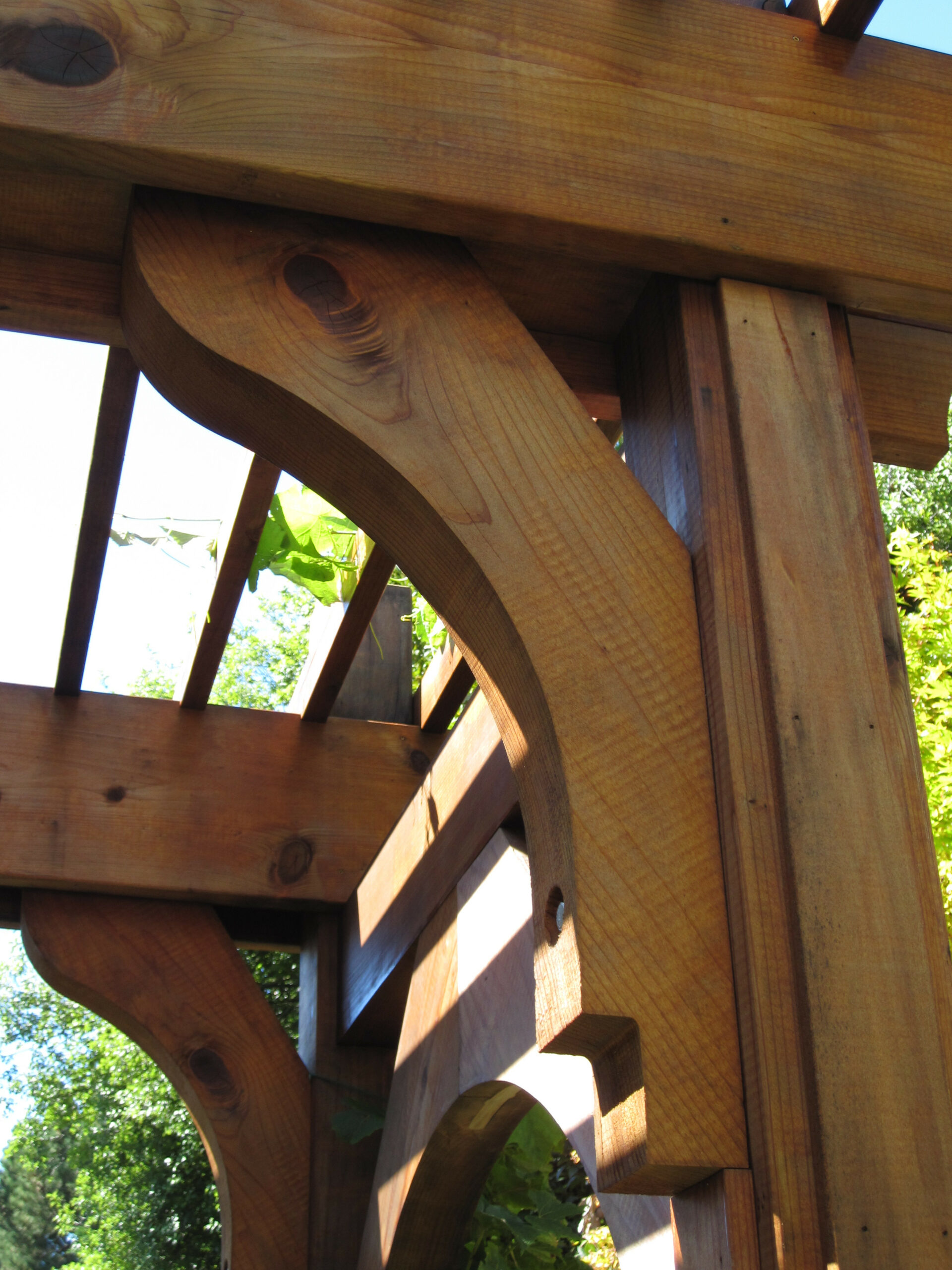 Close-up of a wooden pergola’s structural design, featuring curved support brackets and sturdy overhead beams in a residential yard.