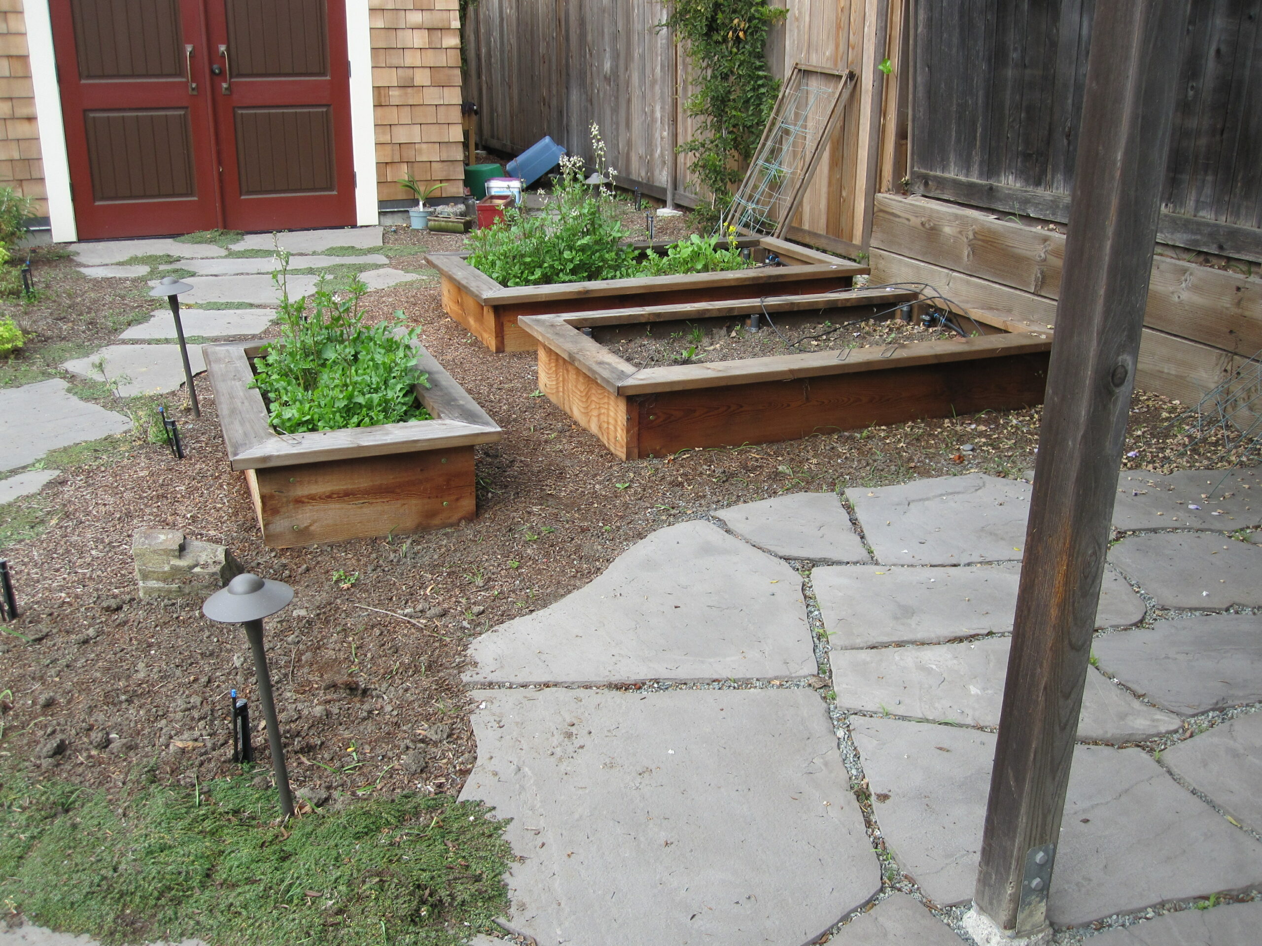 Raised garden beds are positioned along a fenced border next to a stone patio, maximizing efficient use of limited backyard space.