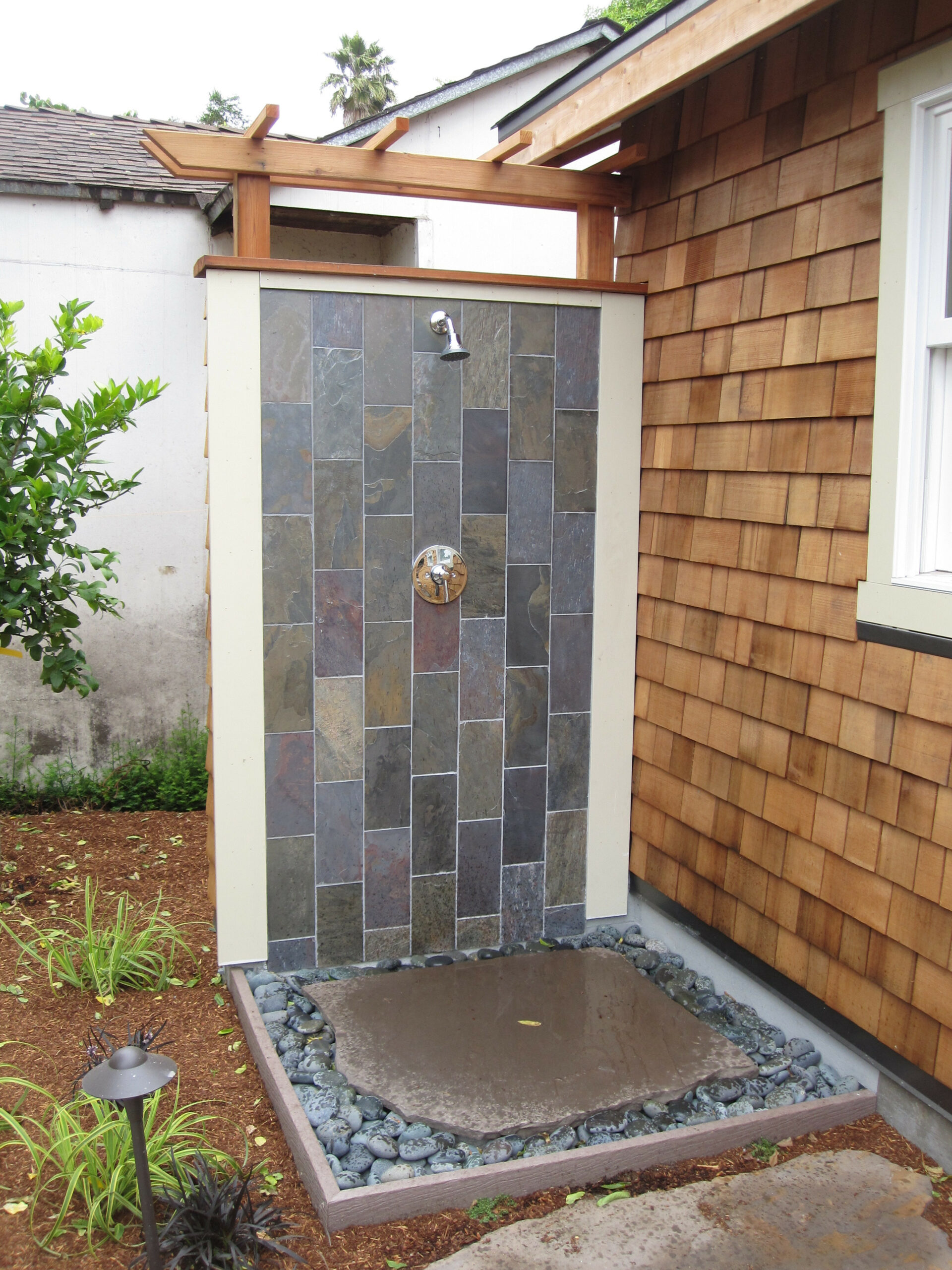 Backyard outdoor shower with tiled wall, single showerhead, durable stone flooring, drainage features, adjacent to wood-sided house.