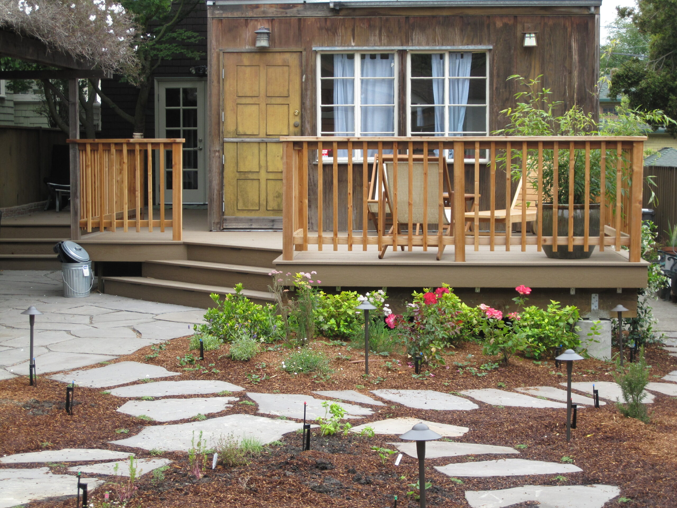 A deck with railings, structurally attached to the house, overlooks a backyard with hardscaped stepping stones and lighting features.
