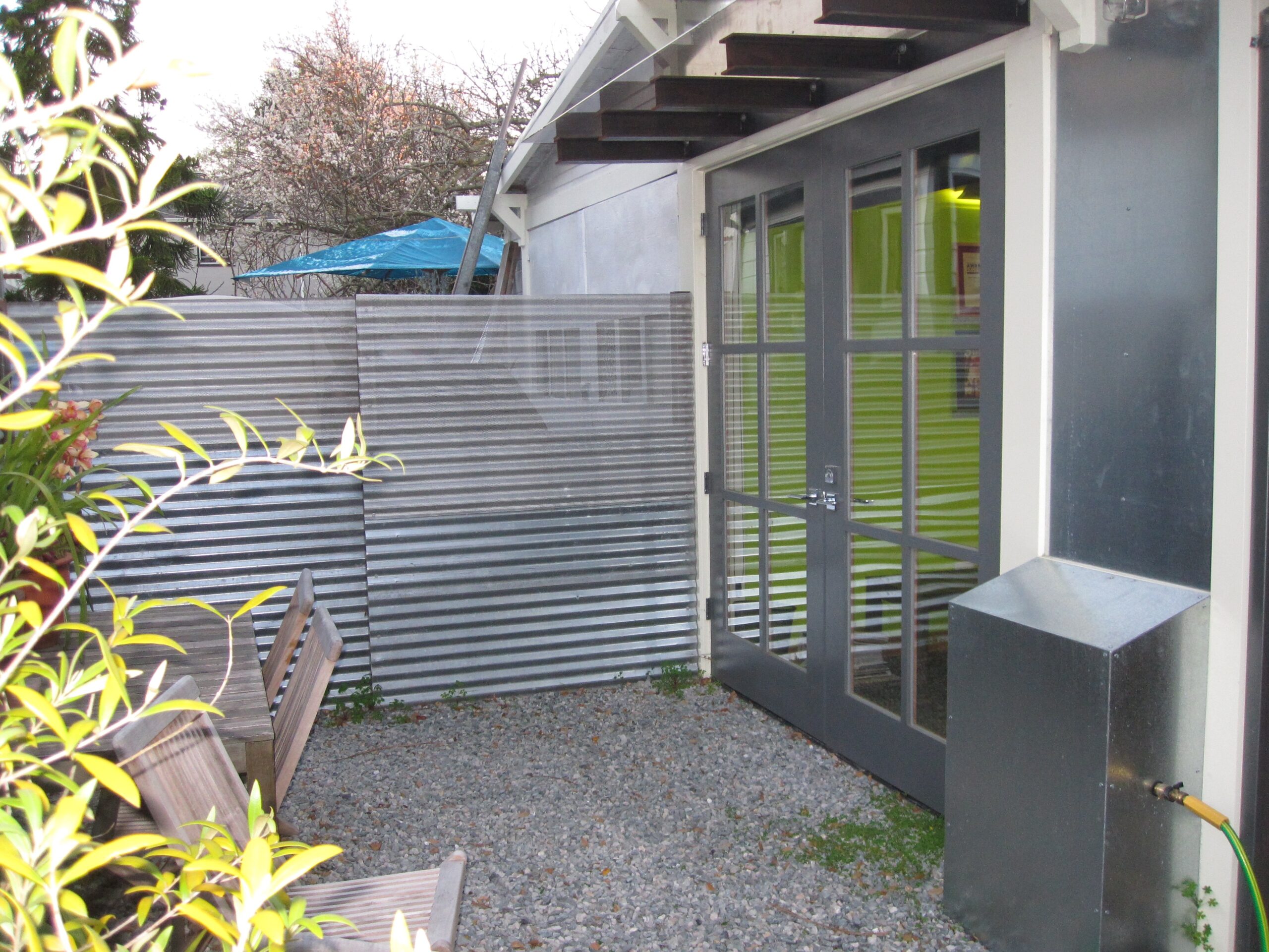 Gravel patio adjacent to corrugated metal fencing, double glass doors provide access to office with visible dividing interior walls.