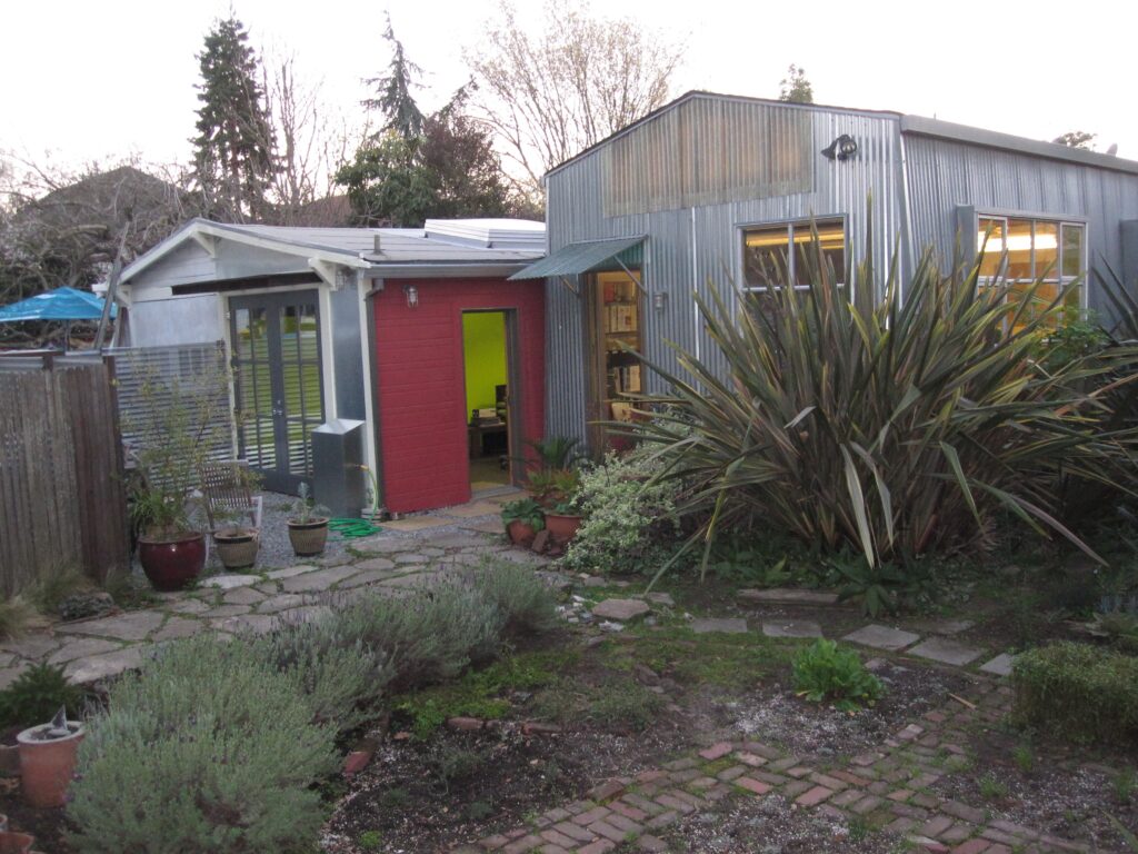 A backyard features a garden and stone path leading to two metal and wood structures, both accessible and showing interior potential.