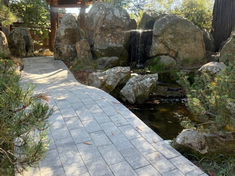 Stone walkway to wooden gate, featuring built-in water elements and large stones set within structured garden landscaping.
