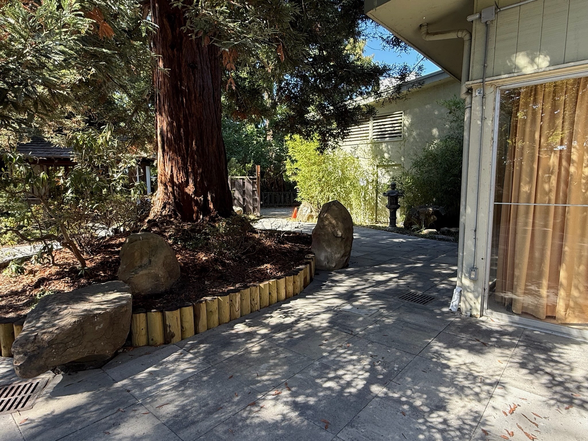 A paved patio with large rocks, mature landscaping, and a building featuring glass doors shows potential for seamless indoor-outdoor flow.