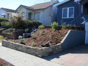 Front yard with sloped, mulched garden bed for drainage, new plantings, and a durable stone retaining wall enhancing structure.