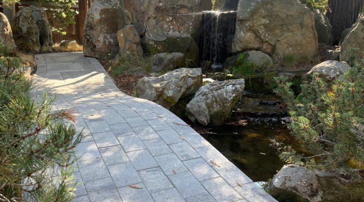 Stone walkway to wooden gate, featuring built-in water elements and large stones set within structured garden landscaping.